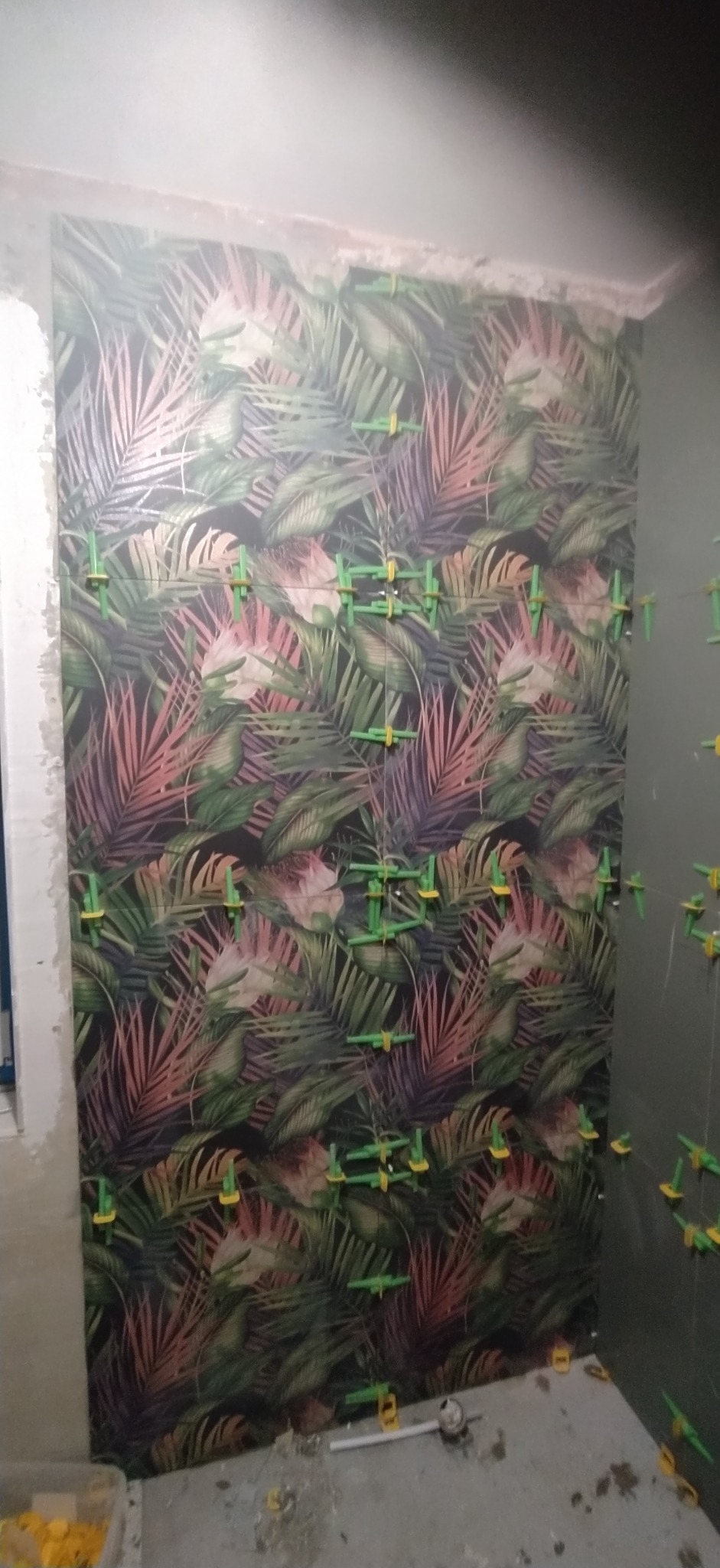 Ściana w trakcie układania płytek z motywem tropikalnych liści, widoczne zielone krzyżyki dystansowe, surowy stan wykończenia.