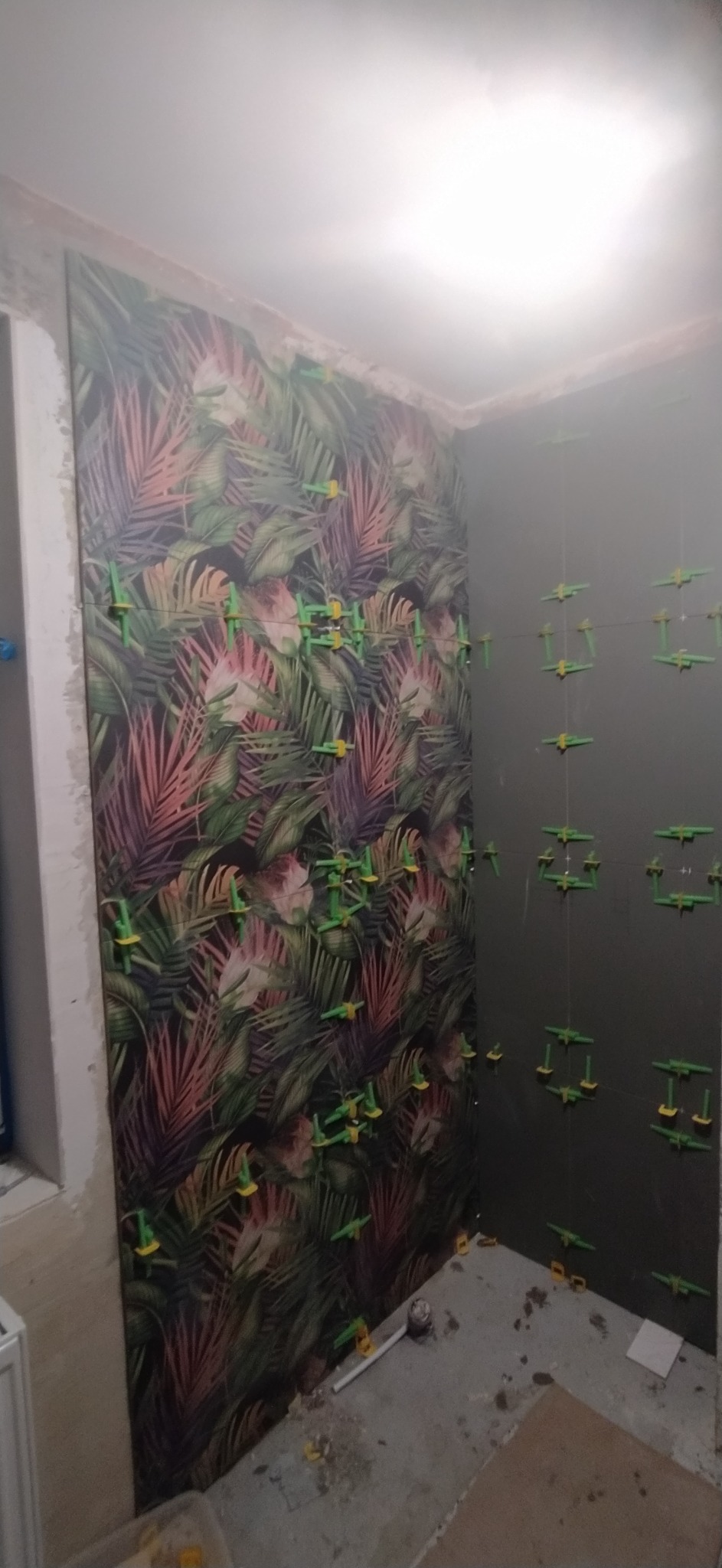 Proces układania płytek z motywem tropikalnych liści na ścianie łazienki, widoczne zielone krzyżyki dystansowe i czarne płytki na sąsiedniej ścianie, surowy stan wykończenia podłogi.