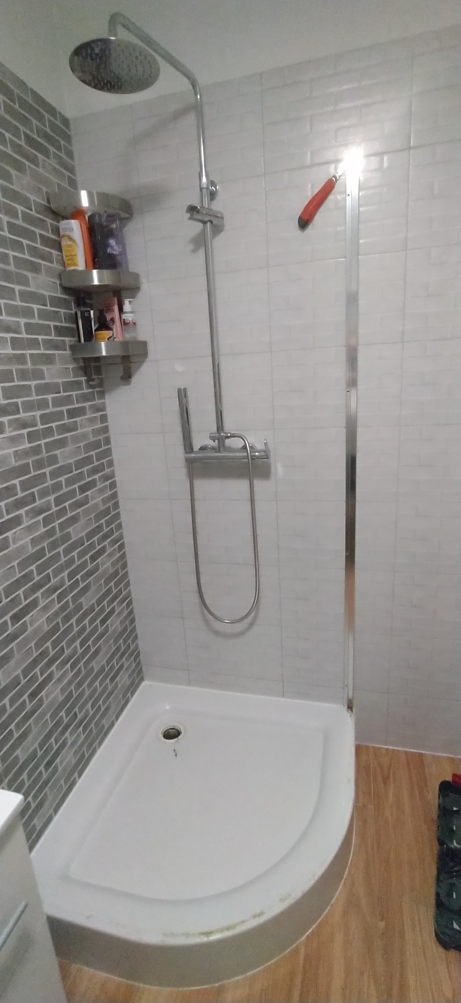 Kabina prysznicowa z deszczownicą i szklanymi drzwiami, ściany wyłożone szarymi i białymi płytkami, półki z kosmetykami po lewej stronie.