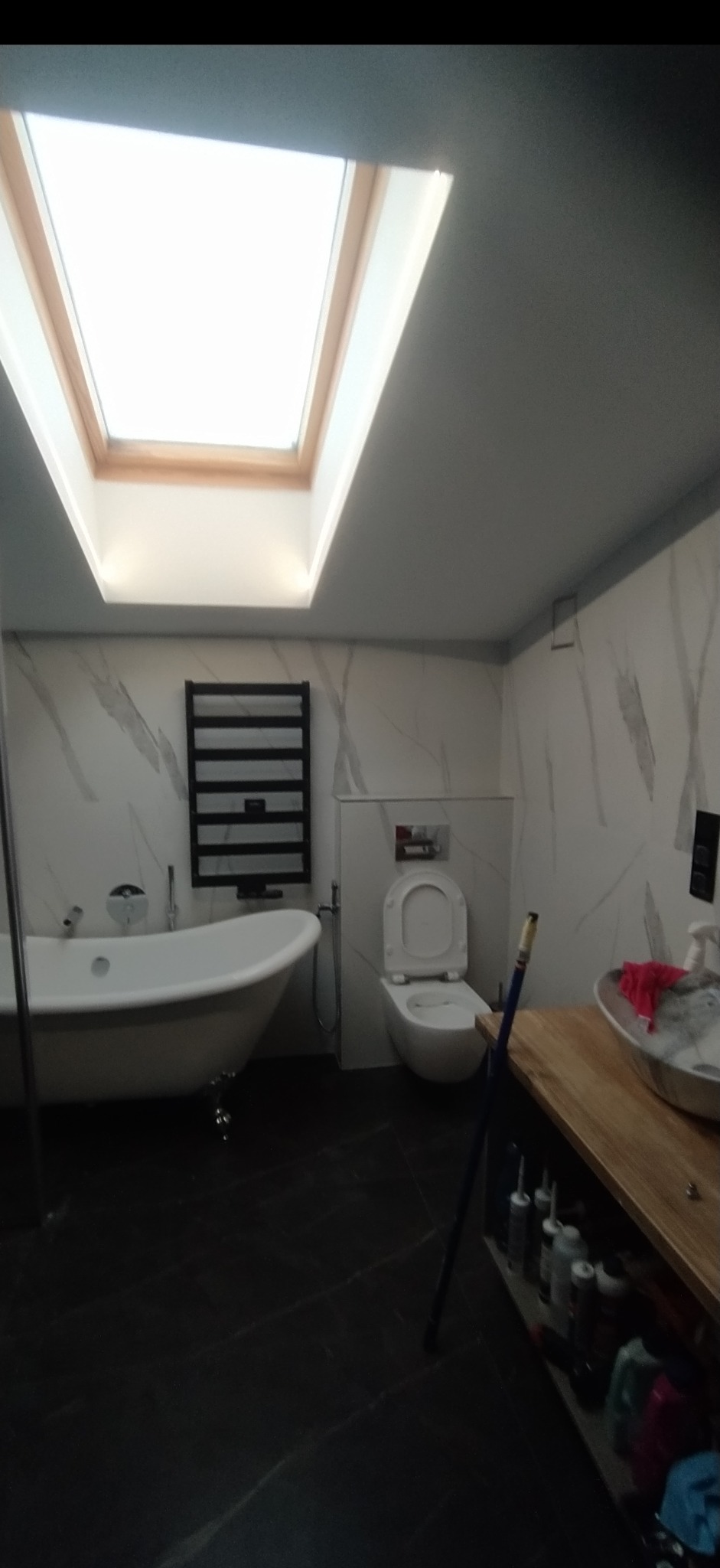 Nowoczesna łazienka z wolnostojącą wanną na lwich nóżkach, grzejnikiem drabinkowym, umywalką na drewnianym blacie i oknem dachowym, wykończona płytkami imitującymi marmur.