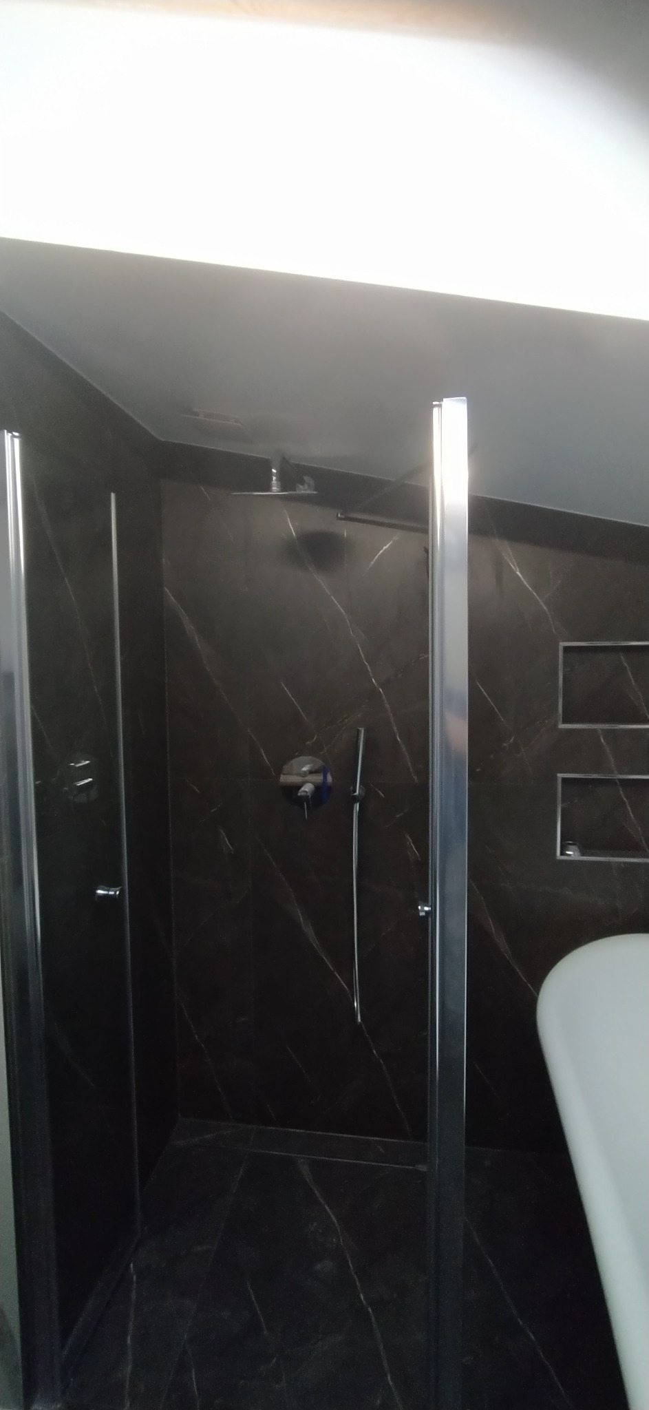 Szklana kabina prysznicowa z czarnymi marmurowymi płytkami, deszczownica i bateria podtynkowa, fragment umywalki widoczny po prawej stronie.