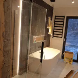 Nowoczesna łazienka z kabiną prysznicową typu walk-in z podświetleniem LED, wolnostojącą białą wanną, marmurową ścianą i czarnym grzejnikiem drabinkowym, widoczny karton po odkurzaczu.