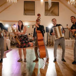 Pięcioosobowy zespół muzyczny grający na scenie podczas wesela: perkusista z bębnem, saksofonistka w kwiecistej spódnicy, kontrabasista, akordeonista i drugi saksofonista w czarnej koszuli z haftem.