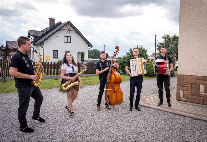 Pięcioosobowy zespół muzyczny z saksofonami, kontrabasem, akordeonem i bębnem pozuje na zewnątrz przed białym domem.