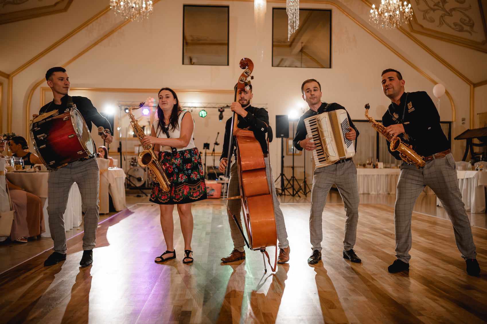 Pięcioosobowy zespół muzyczny grający na scenie podczas wesela: perkusista z bębnem, saksofonistka w kwiecistej spódnicy, kontrabasista, akordeonista i drugi saksofonista w czarnej koszuli z haftem.