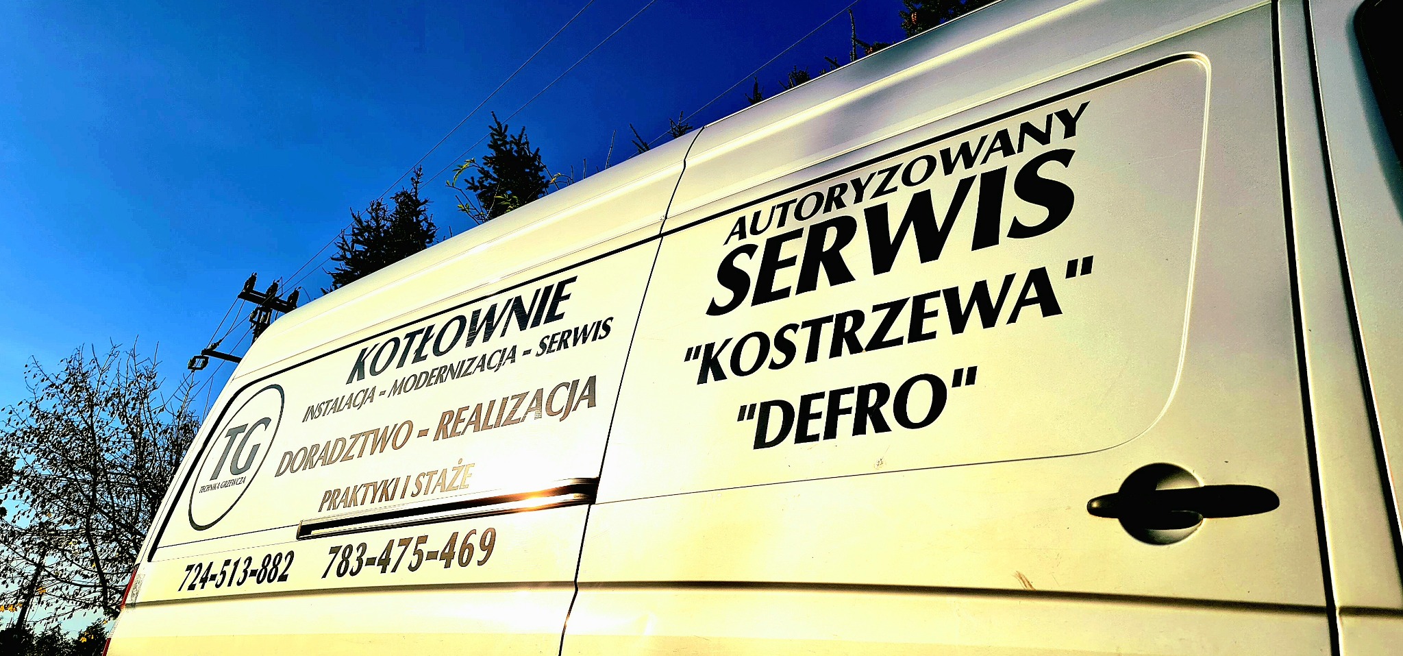 Biały samochód serwisowy z napisem 'Kotłownie: instalacja, modernizacja, serwis. Autoryzowany serwis Kostrzewa, Defro' oraz numerami telefonów, na tle błękitnego nieba.