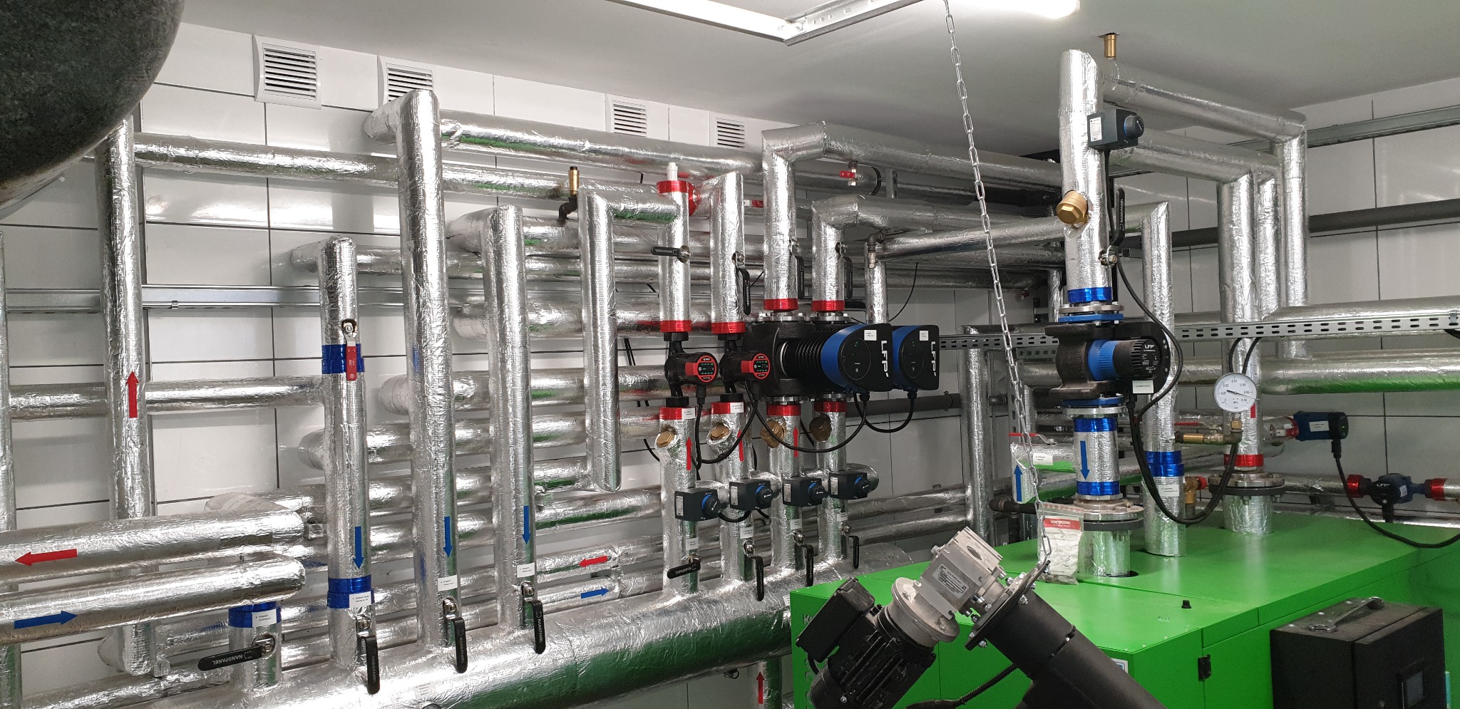 Złożona instalacja grzewcza z rurami w srebrnej izolacji, czerwonymi i niebieskimi zaworami, oraz zielonym kotłem w pomieszczeniu z białymi kafelkami.