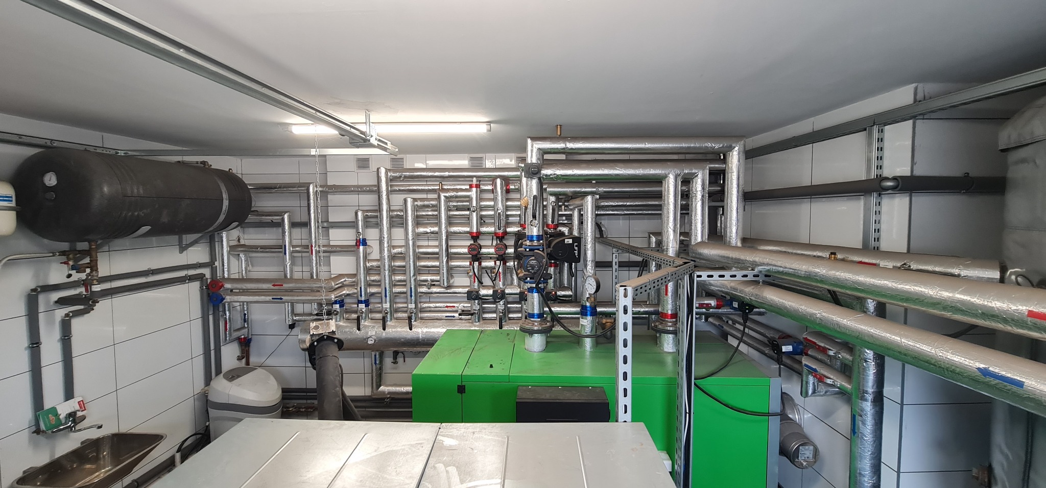 Węzeł cieplny z zielonym piecem, zbiornikiem wyrównawczym i licznymi rurami z izolacją termiczną w pomieszczeniu z białymi płytkami.