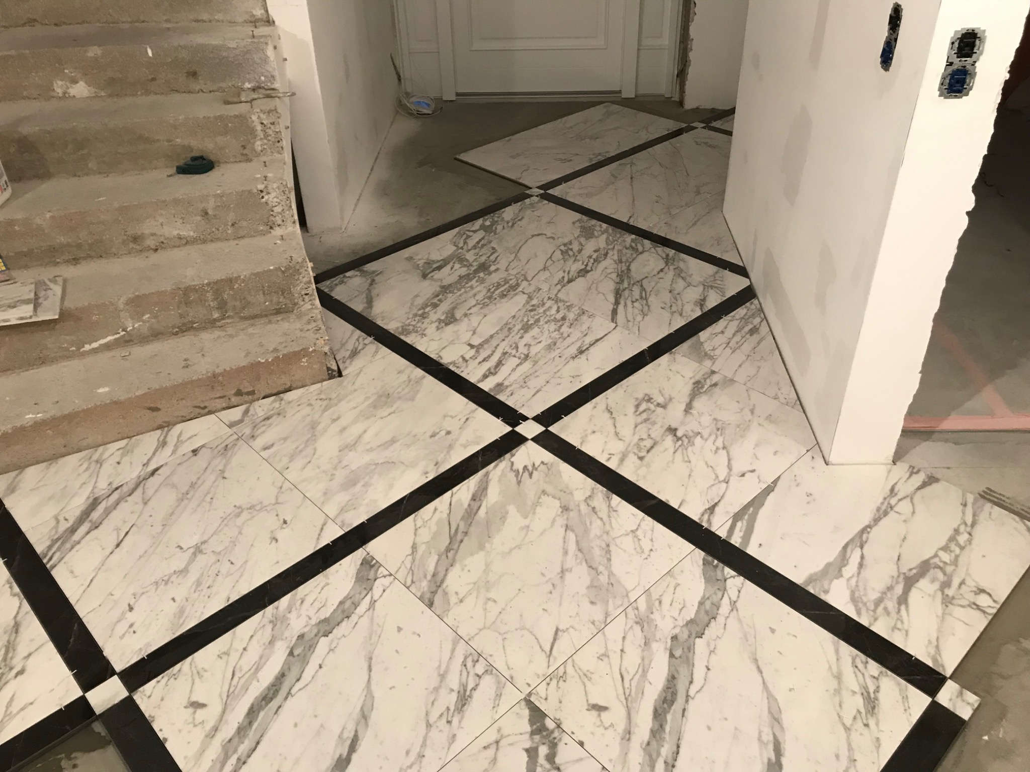 Układanie marmurowej podłogi z czarnymi akcentami w nowym domu, widoczne niedokończone ściany i schody betonowe w tle.