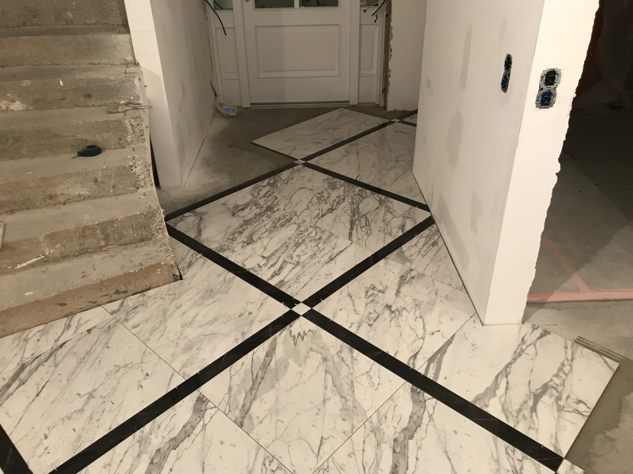 Układanie marmurowej posadzki z czarnymi akcentami w korytarzu, widoczne niedokończone ściany i schody w surowym stanie.