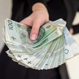 Dłoń kobiety w czarnym ubraniu trzymająca wachlarz banknot&oacute;w polskich złotych o nominałach 100 i 50 zł, skierowany w stronę widza.