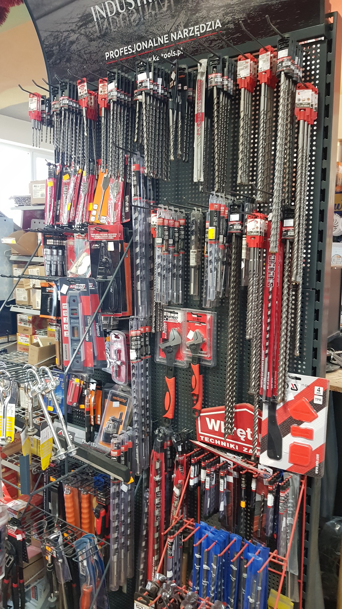 Ekspozycja różnorodnych wierteł i narzędzi ręcznych, w tym kluczy nastawnych, poziomic i śrubokrętów, prezentowanych na metalowym regale w sklepie z narzędziami.