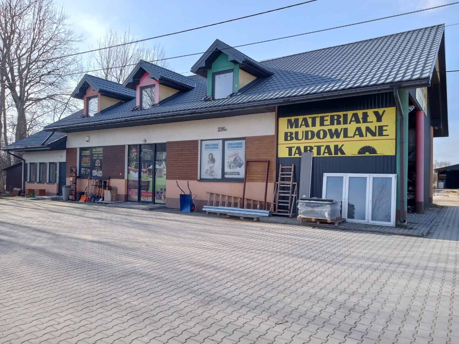 Fasada budynku z szyldem 'Materiały Budowlane Tartak' w Busko-Zdroju, przed budynkiem wystawione okna, drabiny i inne materiały budowlane.