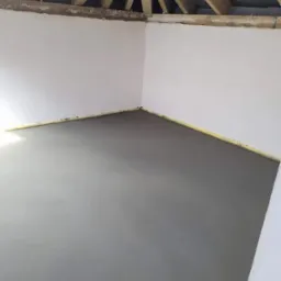 Świeżo wylana, gładka wylewka w rogu pomieszczenia z drewnianą konstrukcją dachu, zabezpieczona żółtą taśmą przy ścianach.