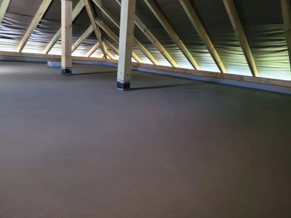 Świeżo wylana, gładka posadzka w surowym stanie poddasza z widoczną drewnianą konstrukcją dachu.