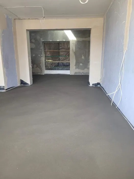 Świeżo wylana, gładka, szara wylewka w pomieszczeniu z przygotowanymi ścianami do malowania i zabezpieczonym otworem okiennym folią.