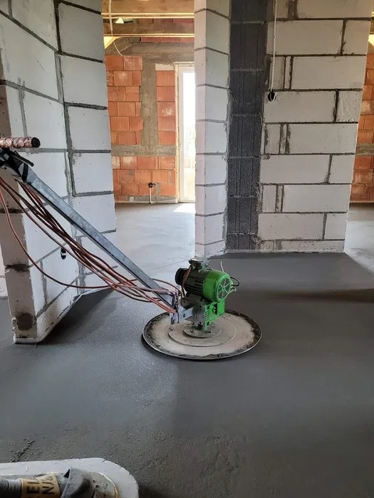 Szlifowanie wylewki betonowej za pomocą maszyny z zielonym silnikiem w surowym wnętrzu budynku z widocznymi pustakami.