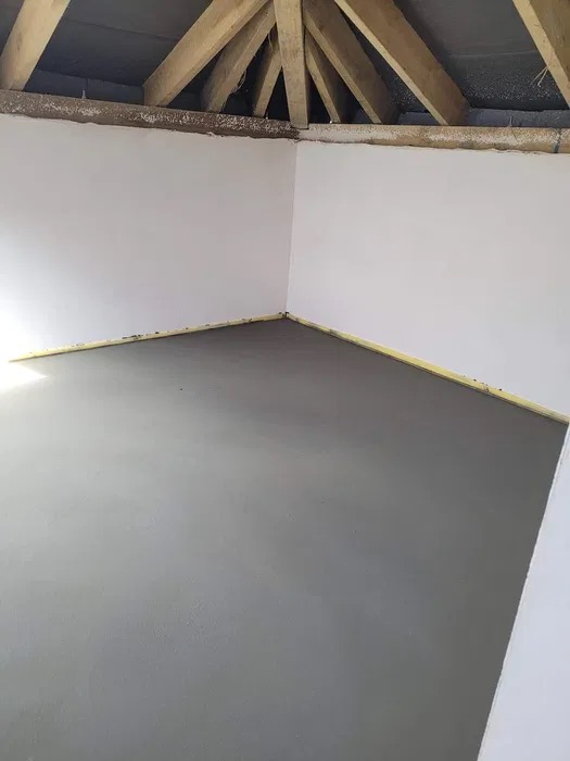 Świeżo wylana, gładka wylewka w rogu pomieszczenia z drewnianą konstrukcją dachu, zabezpieczona żółtą taśmą przy ścianach.