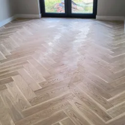 Parkiet ułożony w jodełkę francuską, świeżo po lakierowaniu, widoczny fragment ściany z gładzią i listwami przypodłogowymi.