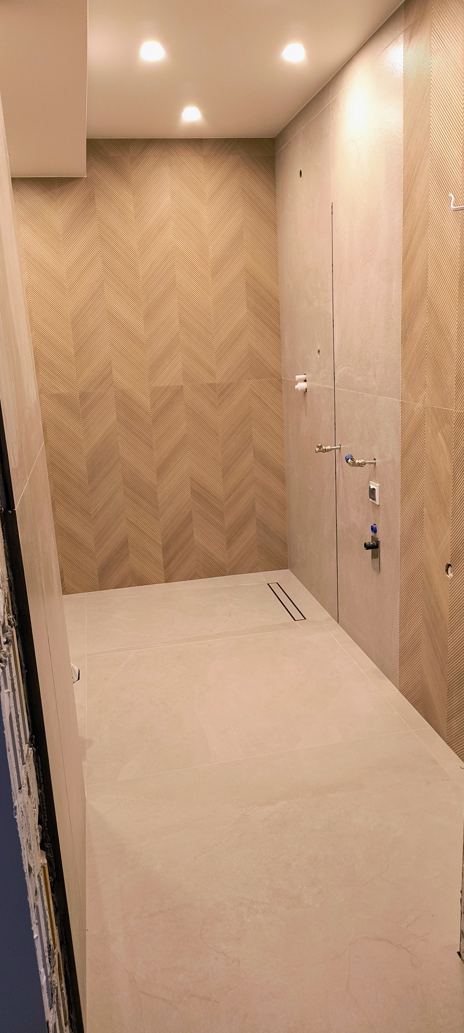 Nowoczesna łazienka w trakcie remontu, z jasnymi płytkami na podłodze i ścianach, wzór jodełki na ścianie, widoczny odpływ liniowy i wystające rury instalacji wodnej.