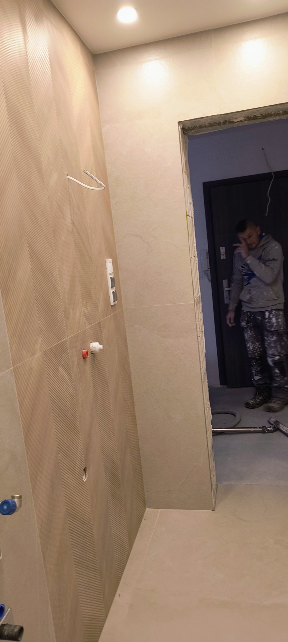 Wykończona ściana łazienki z beżowymi płytkami o wzorze jodełki, widoczne przyłącza wodne i elektryczne. W tle majster w ubraniu roboczym, prawdopodobnie podczas przerwy.