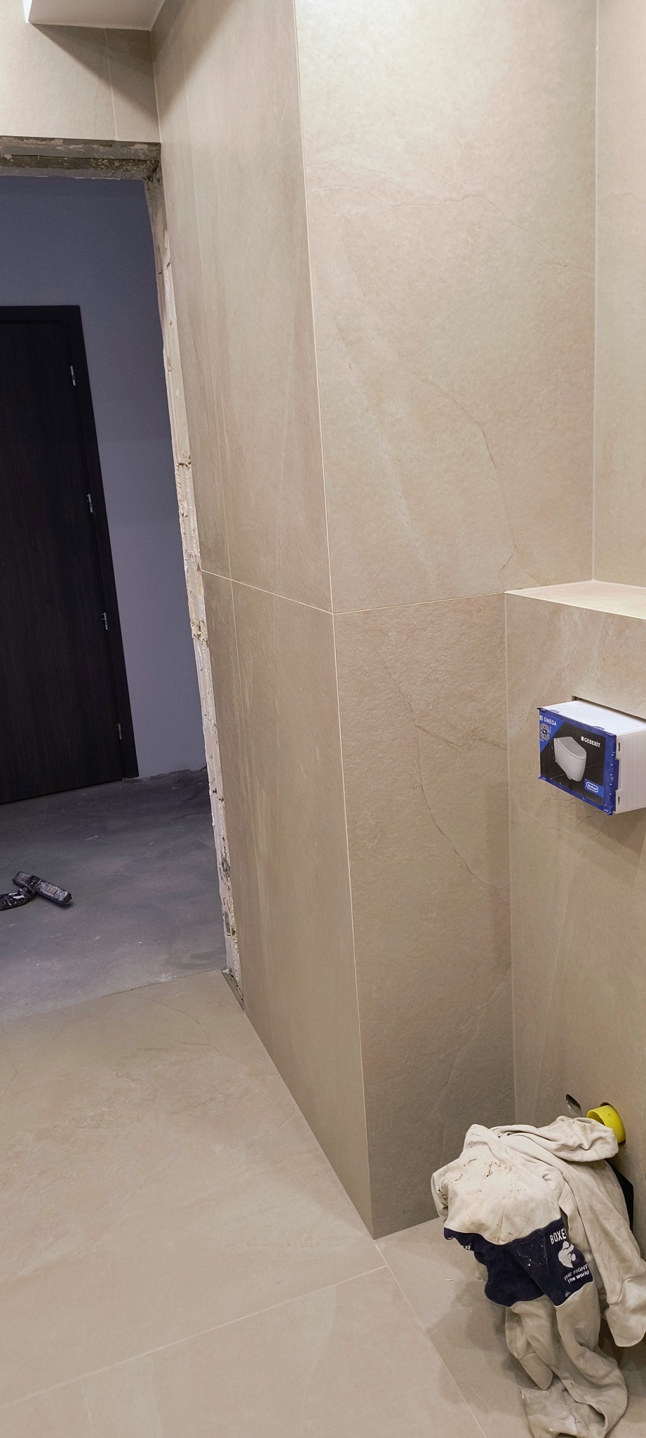 Układanie beżowych płytek w łazience, widoczny fragment ściany z zamontowaną podtynkową instalacją WC marki Geberit, robocze ubranie w rogu i niedokończone obramowanie drzwi.