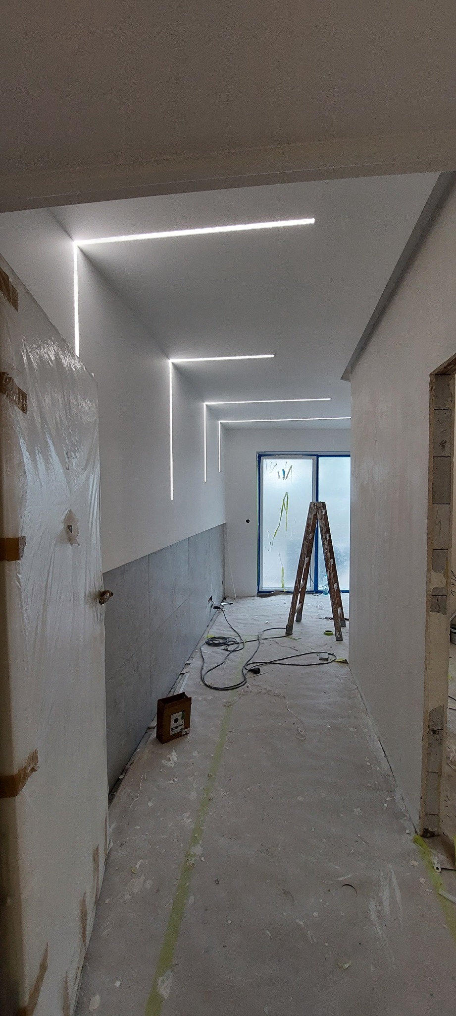 Korytarz w trakcie prac wykończeniowych z zamontowanym oświetleniem liniowym LED na suficie i ścianach, zabezpieczone drzwi folią ochronną, widoczna drabina i narzędzia.