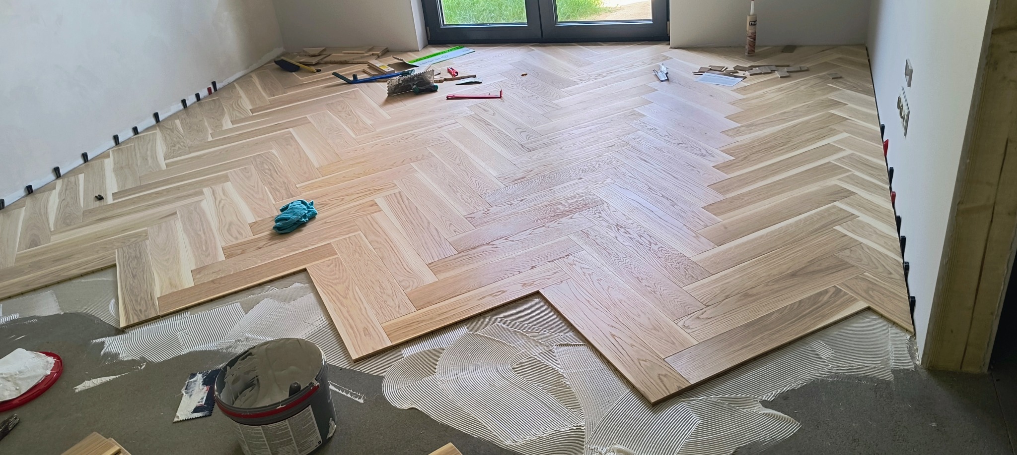Układanie parkietu w jodełkę: widoczne klej, docięte deski, narzędzia i szmatka w pomieszczeniu z oknem.