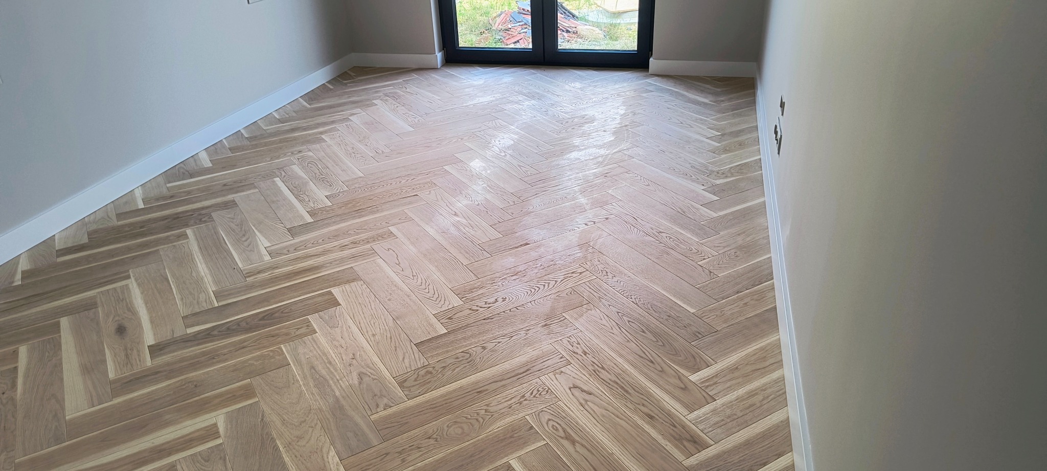 Parkiet ułożony w jodełkę francuską, świeżo po lakierowaniu, widoczny fragment ściany z gładzią i listwami przypodłogowymi.