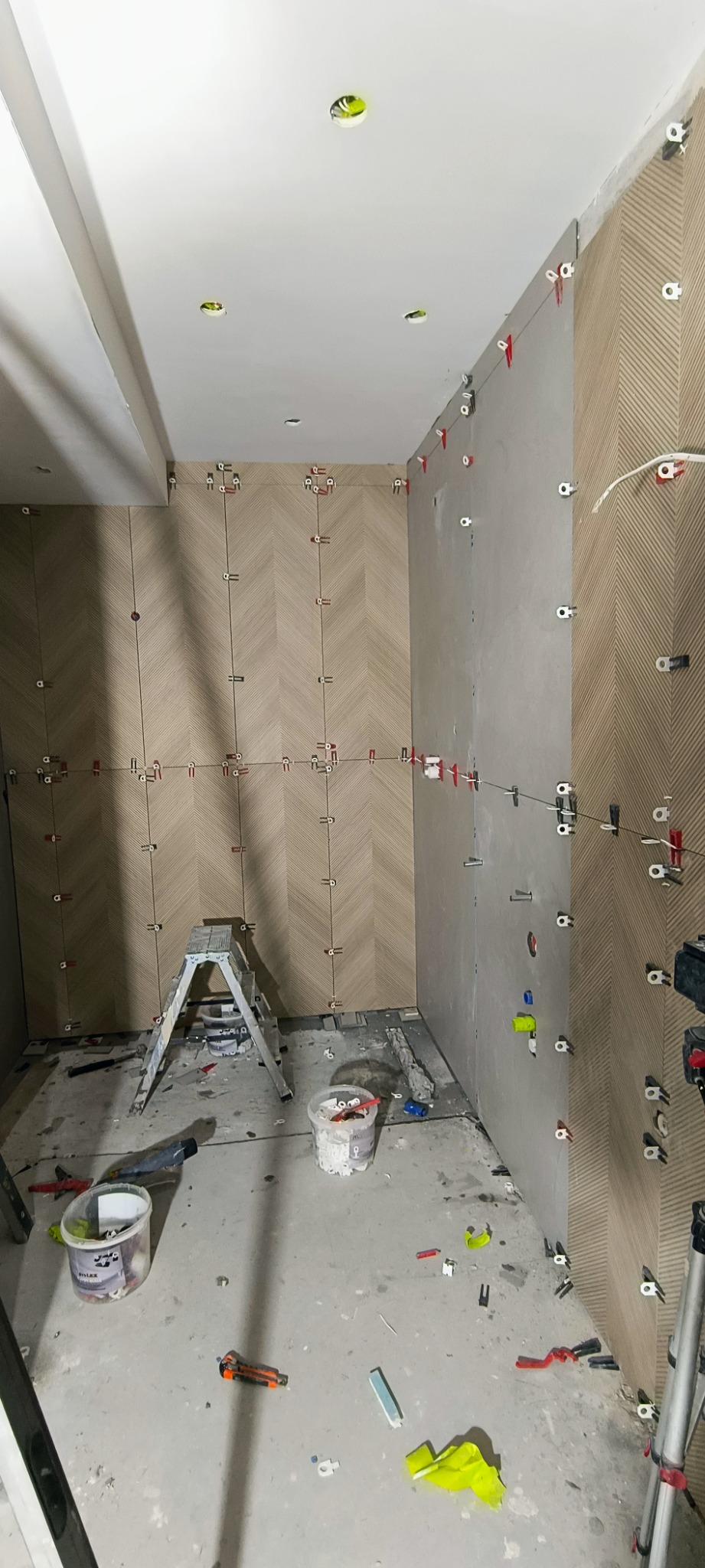 Układanie płytek w jodełkę na ścianie łazienki w trakcie remontu, widoczne poziomice, krzyżyki dystansowe i drabina, podłoga zakurzona z resztkami materiałów budowlanych.
