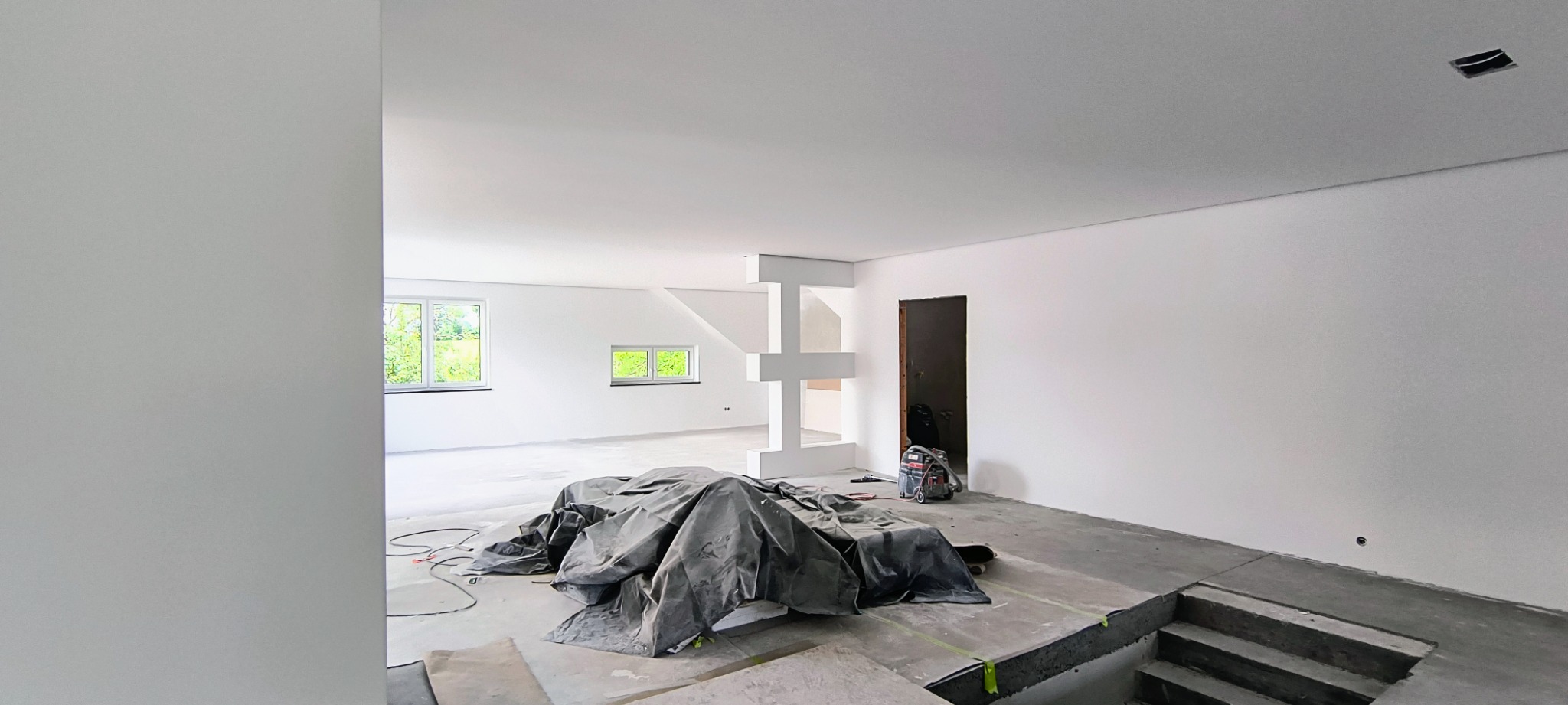 Wnętrze w trakcie remontu: świeżo pomalowane białe ściany, zabezpieczona podłoga, widoczne schody w surowym betonie, okna z widokiem na zieleń i odkurzacz przemysłowy w tle.