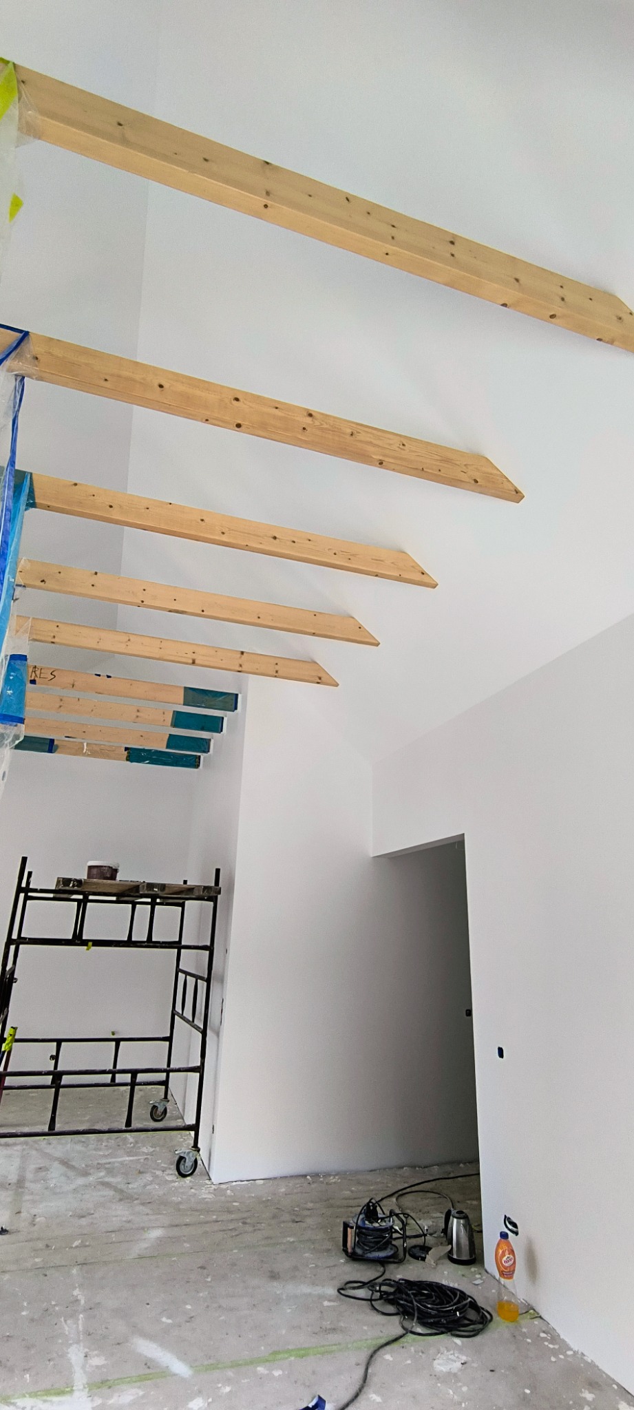 Wnętrze w trakcie prac wykończeniowych: świeżo pomalowane białe ściany, drewniane belki stropowe, rusztowanie i narzędzia malarskie na podłodze.