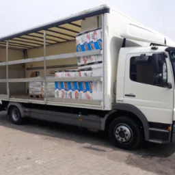 Otwarty, biały samoch&oacute;d ciężarowy z załadowanym materiałem izolacyjnym w opakowaniach z niebieskimi i czerwonymi elementami graficznymi, stojący na brukowanym placu.