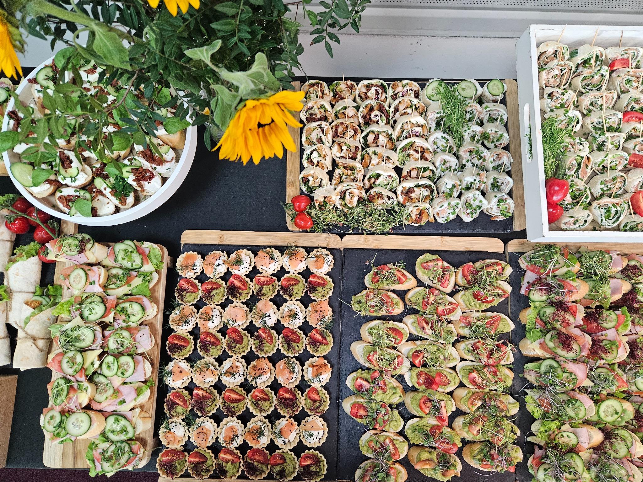 Bogato zastawiony stół z różnorodnymi przekąskami cateringowymi: tartaletki, kanapki, tortille. Dekoracja z kwiatów słonecznika. Idealne na imprezy firmowe i świąteczne.