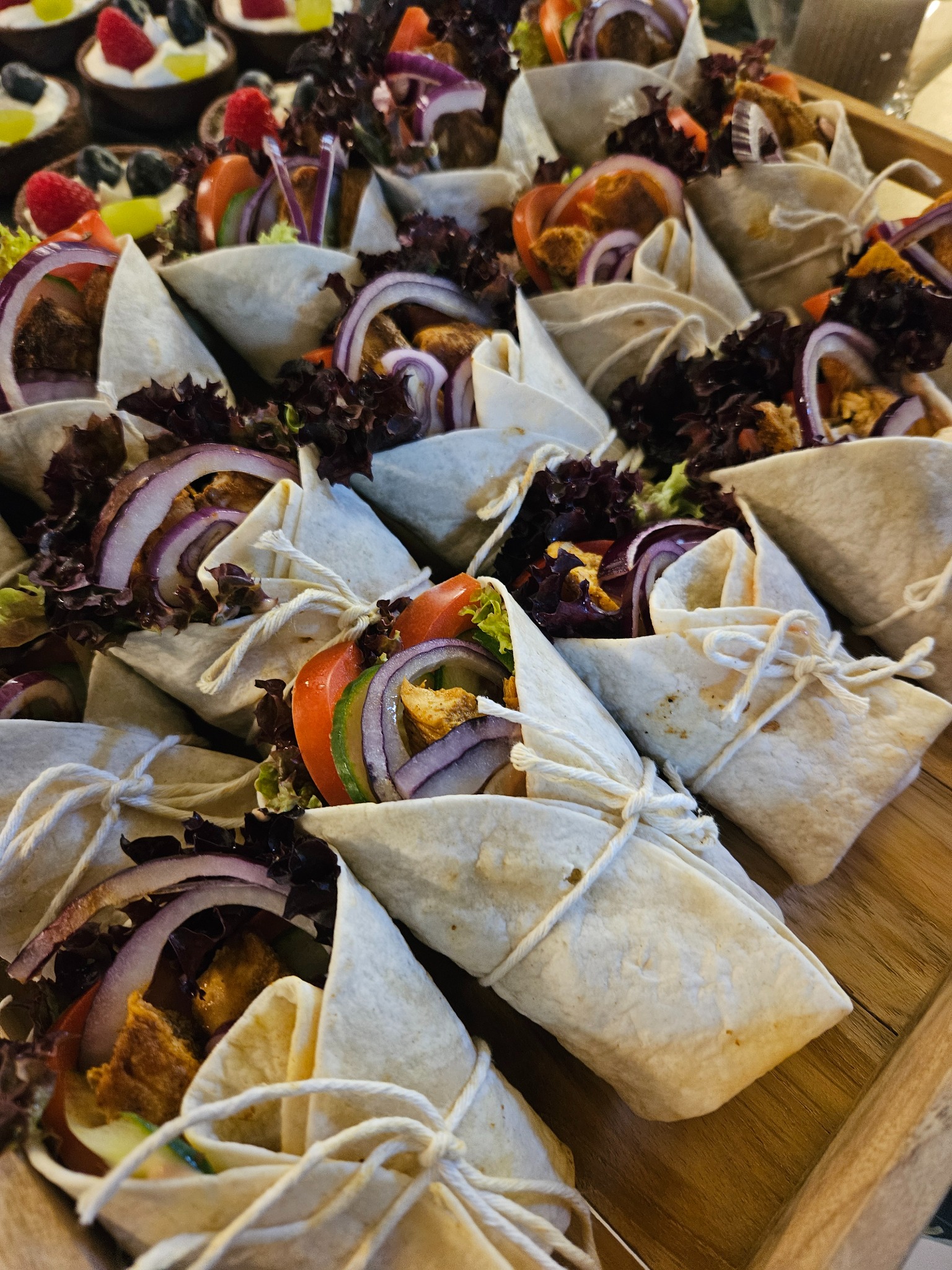 Apetyczne tortille z warzywami i mięsem, zawinięte i związane sznurkiem, ułożone na drewnianej tacy obok mini deserów. Idealne na catering firmowy lub imprezę.