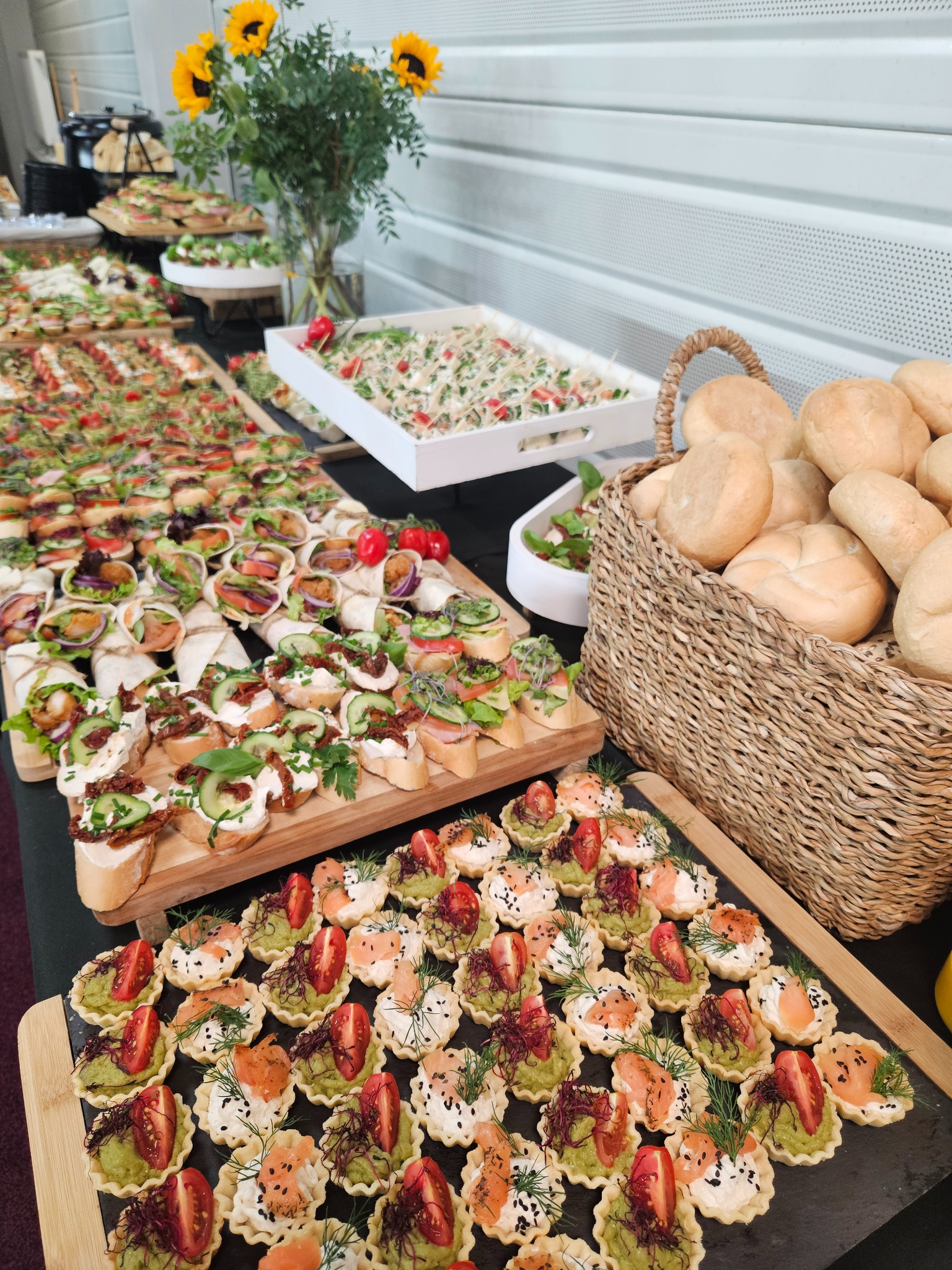 Stół cateringowy z różnorodnymi przekąskami: tartaletki z łososiem i awokado, kanapki, wrapy i sałatki. W tle wazon ze słonecznikami i biała ściana.