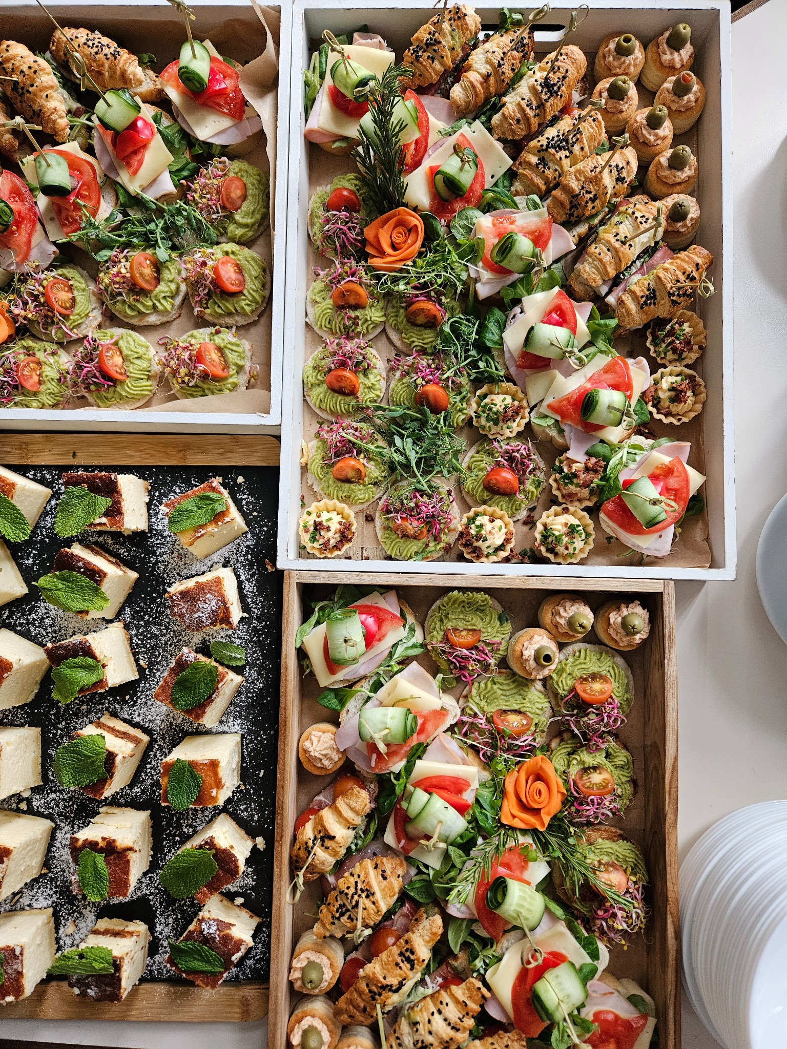 Dekoracyjne przekąski cateringowe: tartinki z awokado, mini-kanapeczki z serem i szynką, croissanty oraz kostki ciasta z miętą, ułożone w drewnianych skrzynkach.