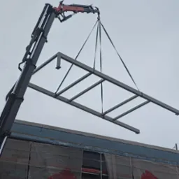 Żuraw podnosi stalową konstrukcję ramową na placu budowy, widoczne rusztowania z osłoną i fragment budynku w tle.
