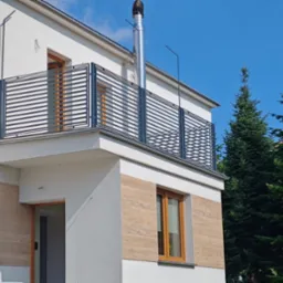 Nowoczesna balustrada balkonowa z poziomych profili metalowych, zamontowana w nowym domu z jasną elewacją i elementami dekoracyjnymi imitującymi drewno.