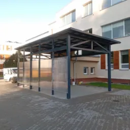 Zadaszenie metalowe z przezroczystymi panelami, widok z zewnątrz, na tle budynku. Zadaszenie na parkingu dla samochodów dostawczych, częściowo widoczny bus.