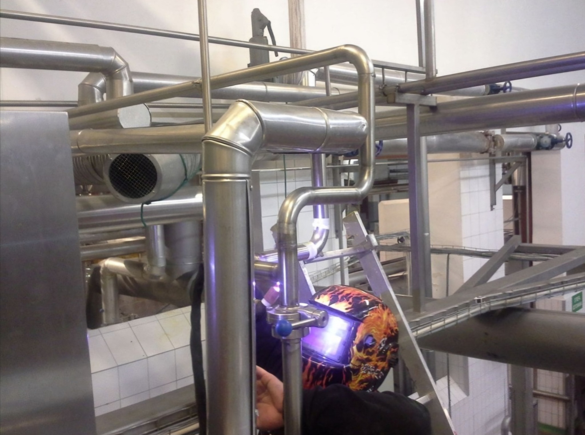 Prace spawalnicze przy rurociągu ze stali nierdzewnej, widoczna spawarka w masce ochronnej z motywem płomieni, drabina i fragmenty instalacji w tle.