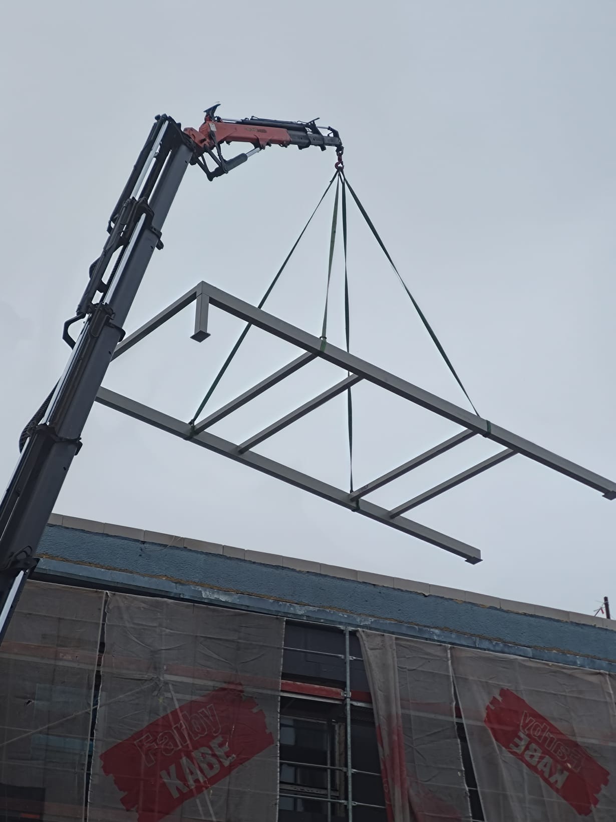 Żuraw podnosi stalową konstrukcję ramową na placu budowy, widoczne rusztowania z osłoną i fragment budynku w tle.