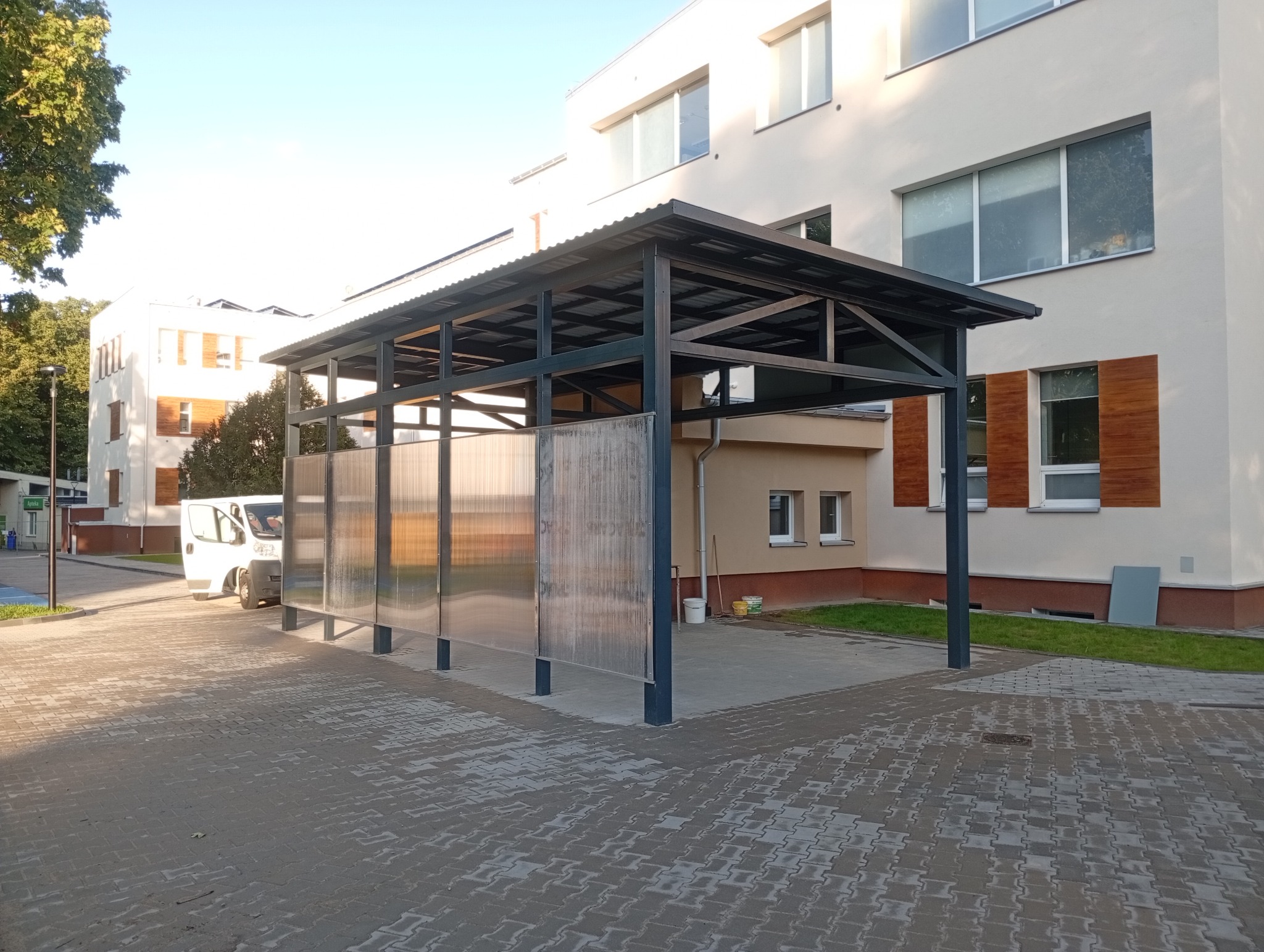 Zadaszenie metalowe z przezroczystymi panelami, widok z zewnątrz, na tle budynku. Zadaszenie na parkingu dla samochodów dostawczych, częściowo widoczny bus.