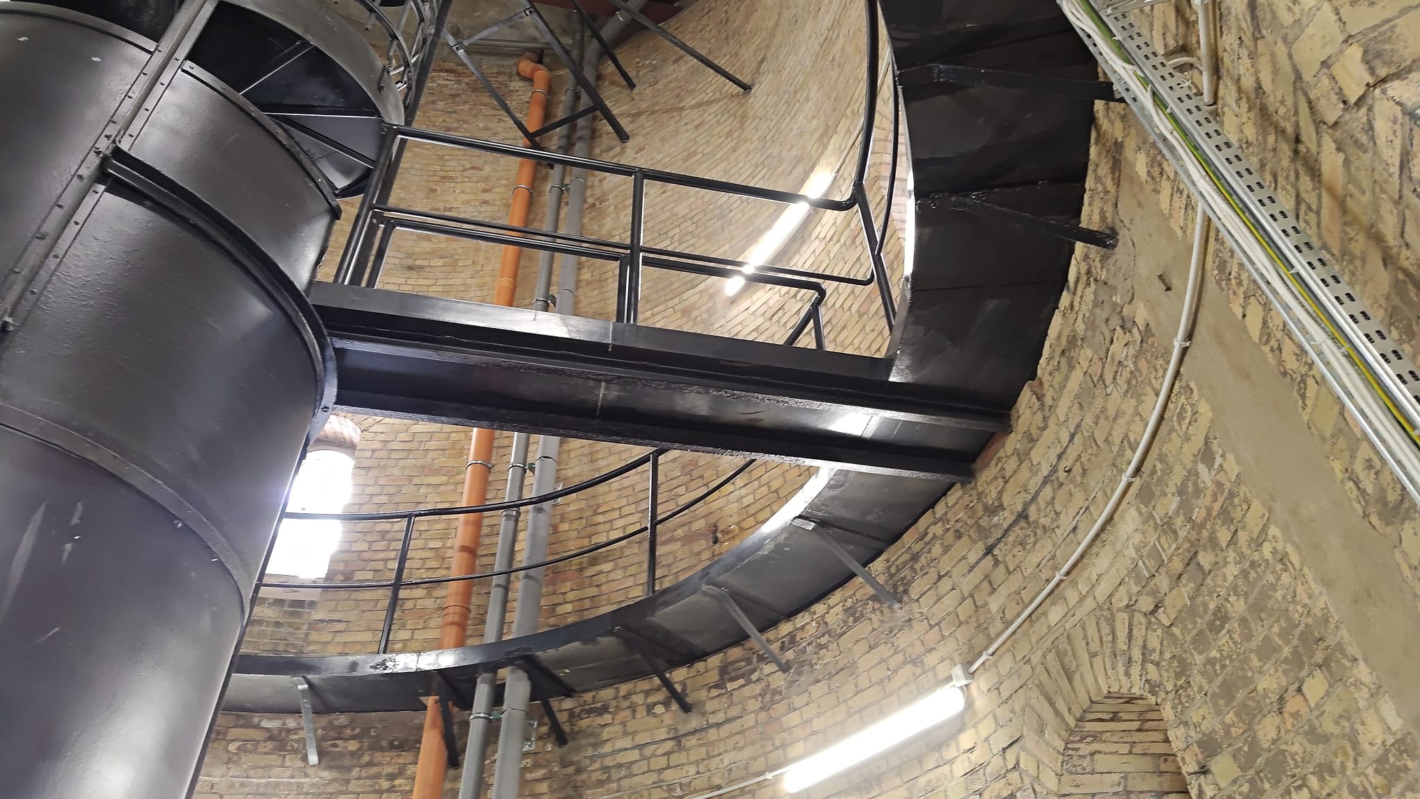 Metalowe schody spiralne wewnątrz ceglanej wieży, z widocznymi instalacjami i oknem w tle. Ujęcie z dołu, podkreślające konstrukcję i jej umiejscowienie.