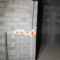 EL-ROB - Instalacja elektryczna w surowym wnętrzu: przewody w ścianie z cegły, pomarańczowe puszki elektryczne, zwój kabli na górze. Widok na podłogę i sufit w stanie deweloperskim.