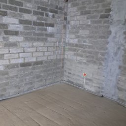 EL-ROB - Surowy pokój z instalacją elektryczną w rurkach przy podłodze i pionową rurką miedzianą, surowe ściany z cegły betonowej i betonowy sufit.