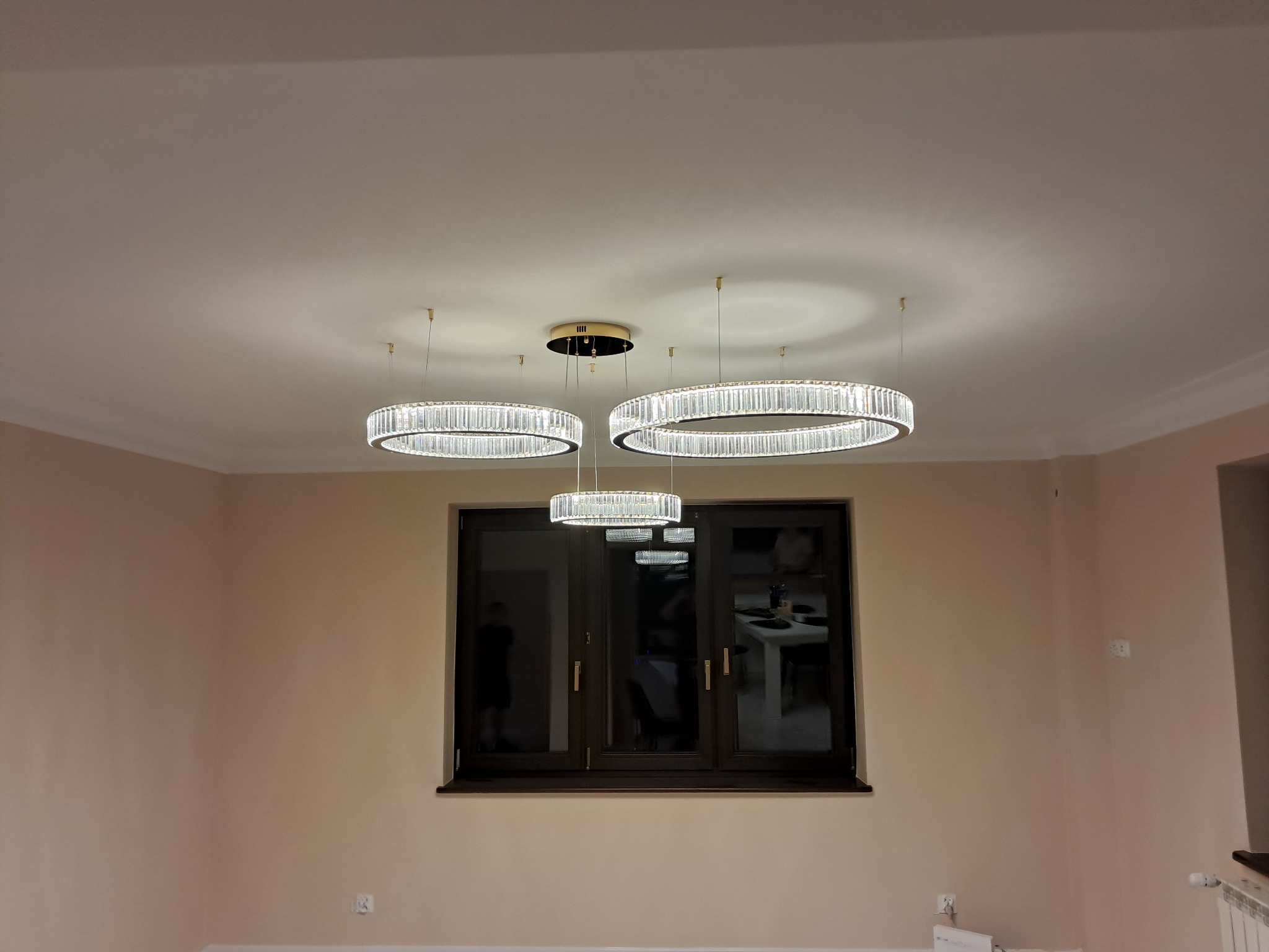 Nowoczesne, okrągłe lampy sufitowe z kryształkami, centralnie umieszczone, symetrycznie rozmieszczone względem okna. Ściany w odcieniach beżu.