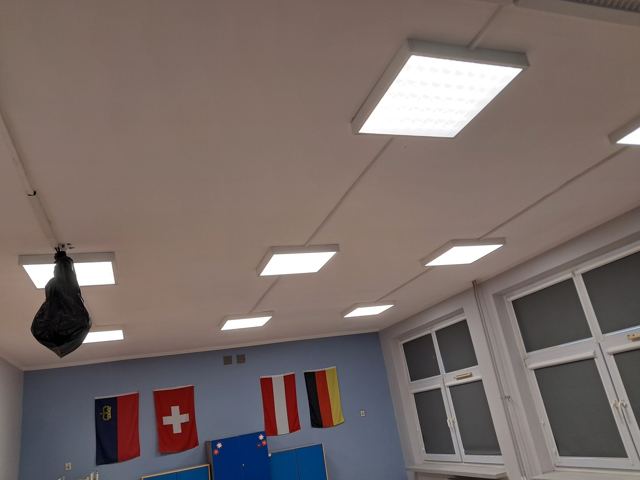 Białe, prostokątne lampy sufitowe w rzędach oświetlają klasę z flagami na ścianach i oknami po prawej. Jedna lampa z workiem na śmieci.