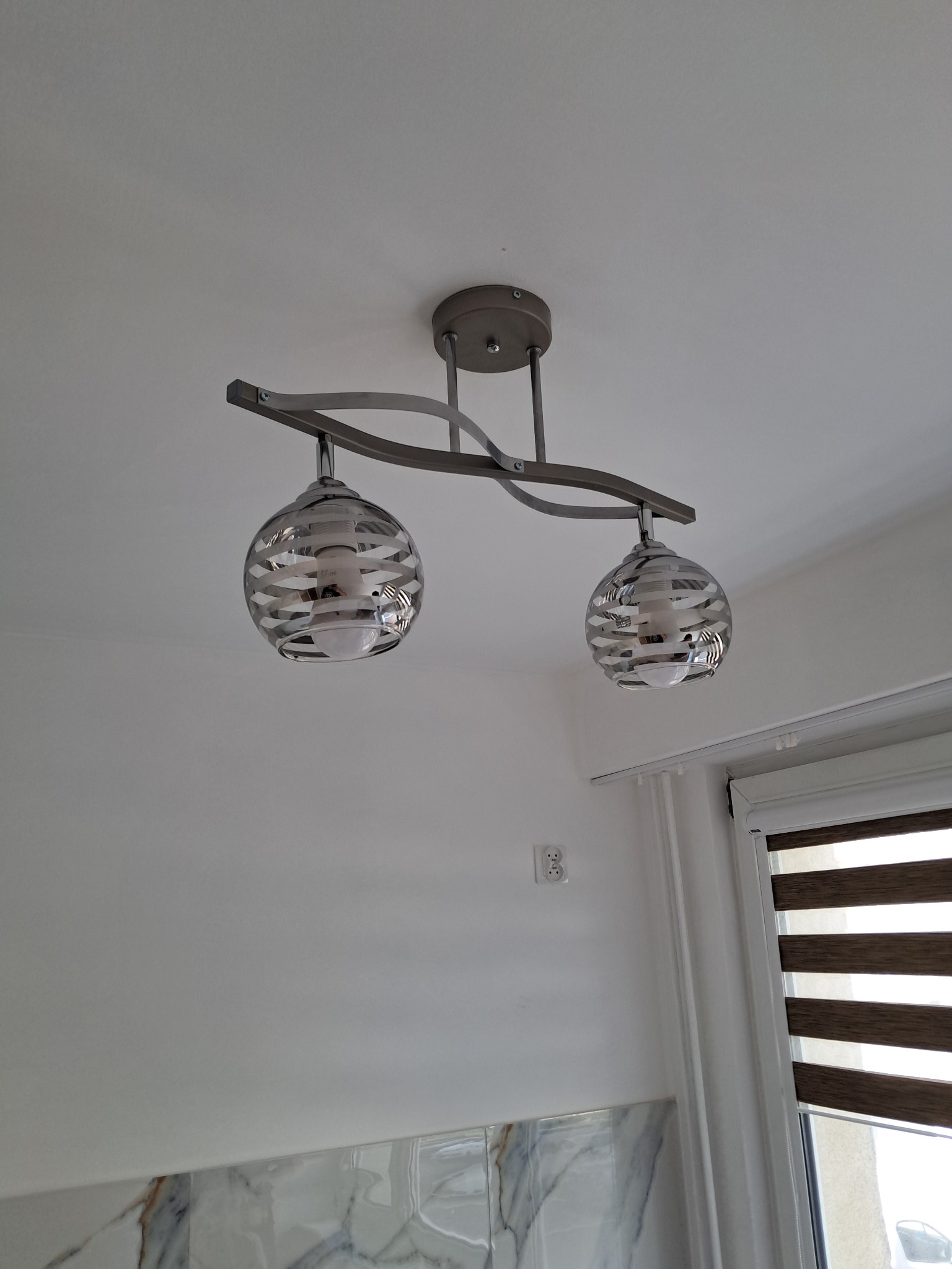 Dwupunktowa lampa sufitowa z dekoracyjnymi, szklanymi kloszami w kształcie kuli, zamontowana na białym suficie w nowoczesnym wnętrzu. Widoczne okno z żaluzjami.