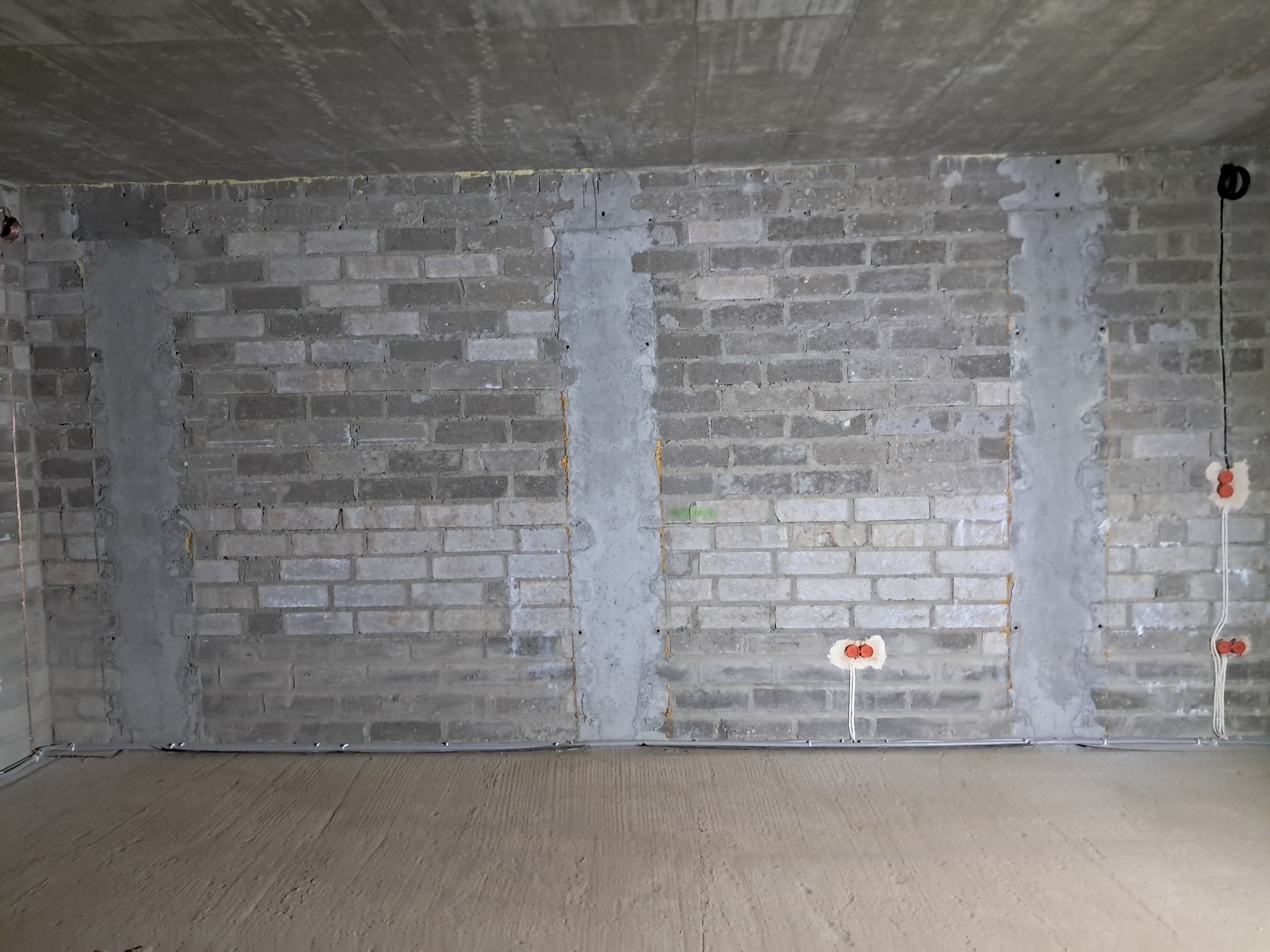 Surowy mur z instalacją elektryczną w bruzdach, widoczne kable i puszki. W tle betonowy sufit. Przewody ułożone wzdłuż ściany. Etap prac wykończeniowych.